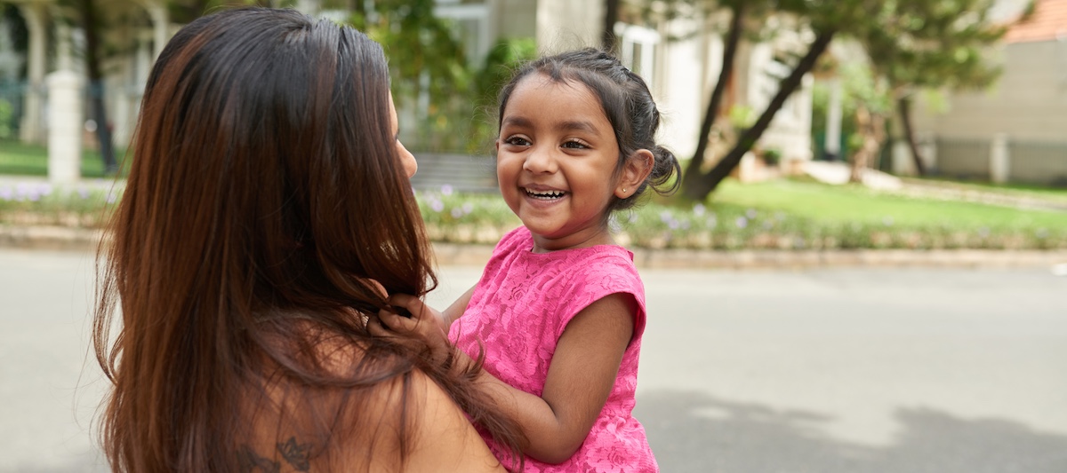 Cheerful little Indian girl looking away with toothy smile while having fun with her mother in front of their house