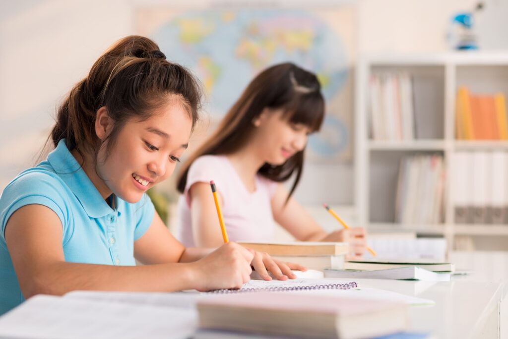 Profile view of pretty teenage student sitting at desk and writing essay with enthusiasm, interior of classroom on background